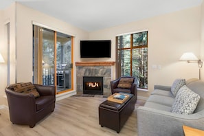 A modern living room with a fireplace, leather sofas, a flat-screen TV, and large windows offering a view of trees.