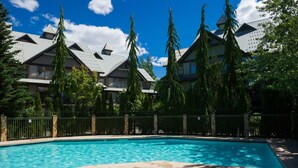 Una piscina con borde de azulejos, rodeada de una cerca, y edificios con techos a dos aguas al fondo.