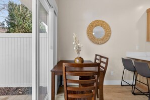 Un comedor con una mesa de madera, dos sillas, un jarrón con flores secas y un espejo tejido en la pared.