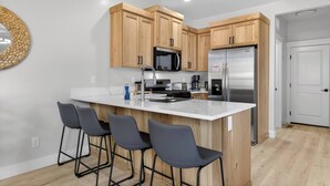 A modern kitchen with wooden cabinets, a stainless steel refrigerator, a microwave, and a bar area with stools. - Nampa`s New Nest -The Renaissance (Nampa)