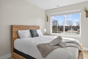 Un dormitorio con cabecera de madera, un ventanal con vistas a una zona residencial y una mesita de noche con lámpara.