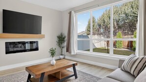 A modern living room with a large window, a flat-screen TV mounted on the wall, a wooden coffee table, and a grey sofa. - Nampa`s New Nest -The Renaissance (Nampa)