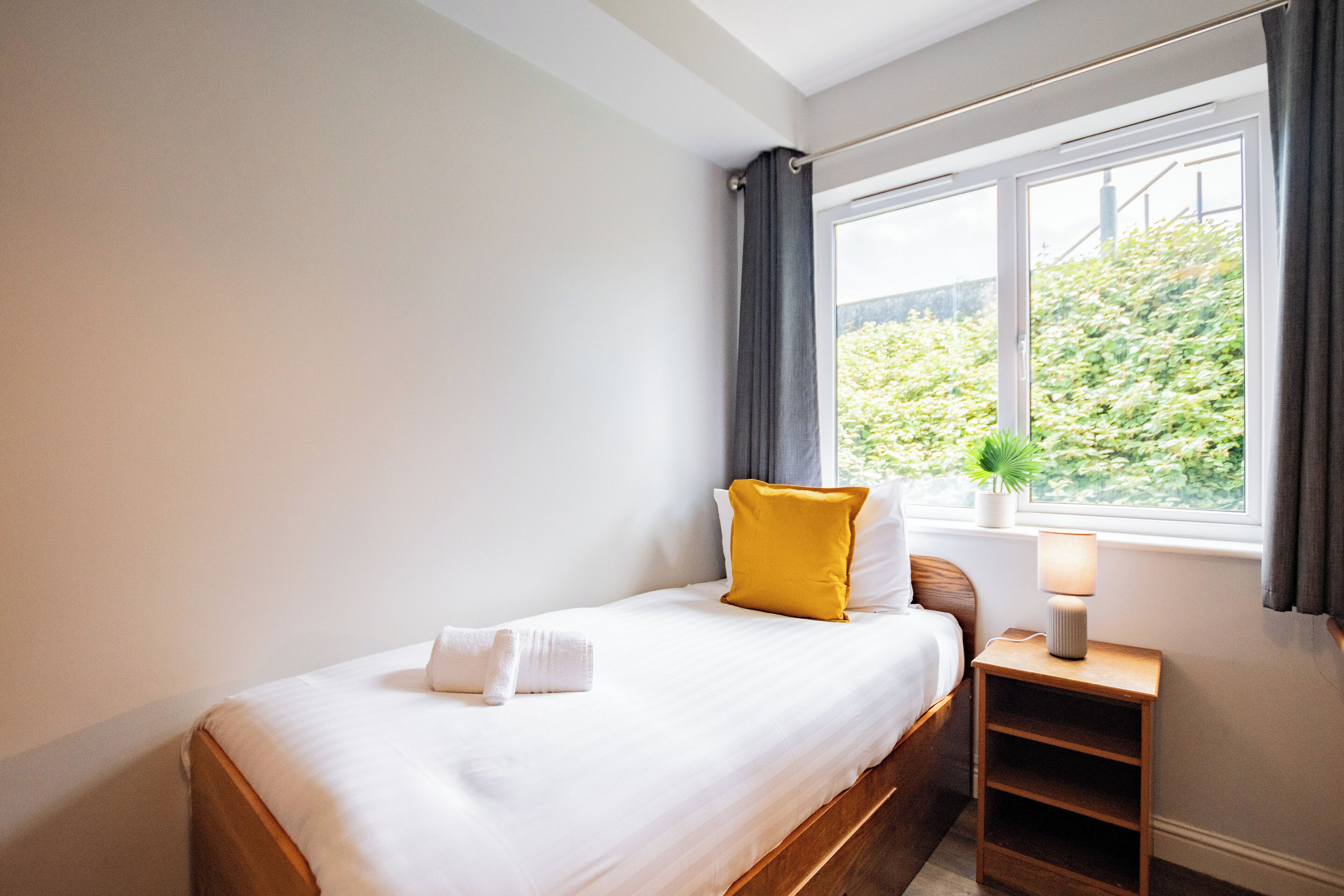 Una cama bien hecha con sábanas blancas y una sola almohada amarilla, una mesita de noche con una lámpara y una ventana con vistas a la vegetación.