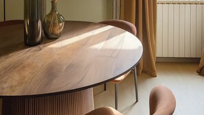 Een moderne eethoek met een ronde tafel, twee stoelen, een vaas met gedroogde planten, een aan de muur bevestigde televisie en een raam met gordijnen.