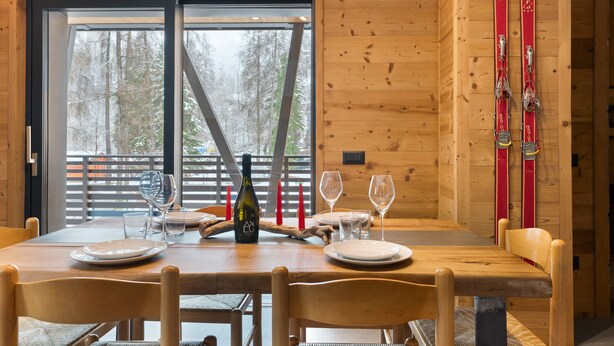 Une table de salle à manger en bois, préparée pour quatre personnes, avec des chaises, une bouteille et des verres placés sur une table près de la fenêtre.