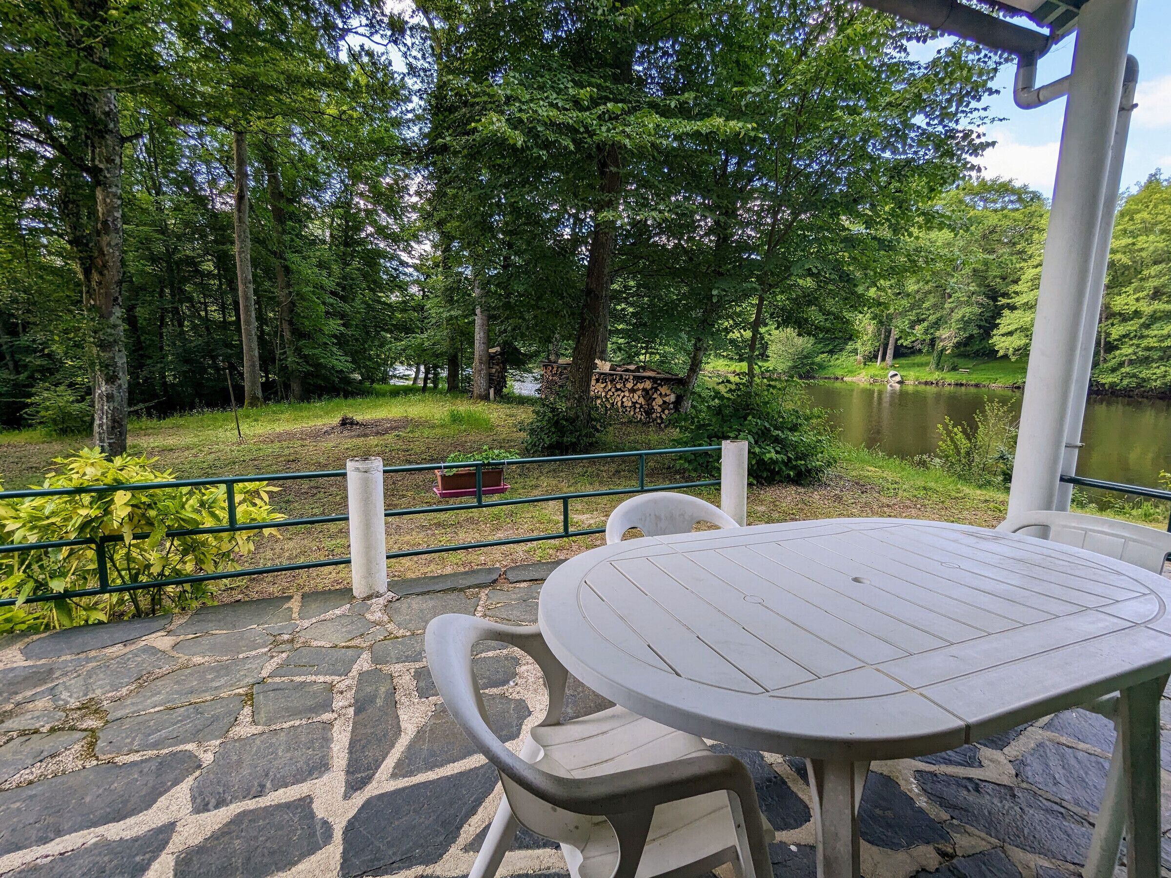 A stone patio with a round table and chairs, overlooking a lake and surrounded by trees.