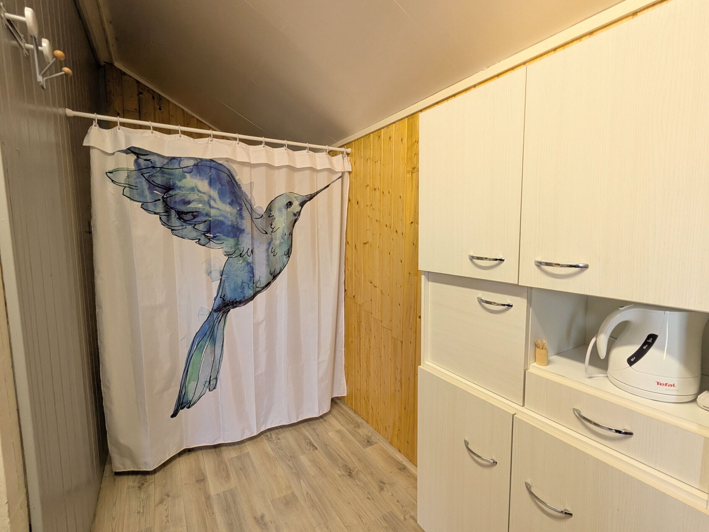 Un baño con cortina de ducha decorada con un diseño de colibrí, paredes de madera y un mueble blanco con cajones y un hervidor.