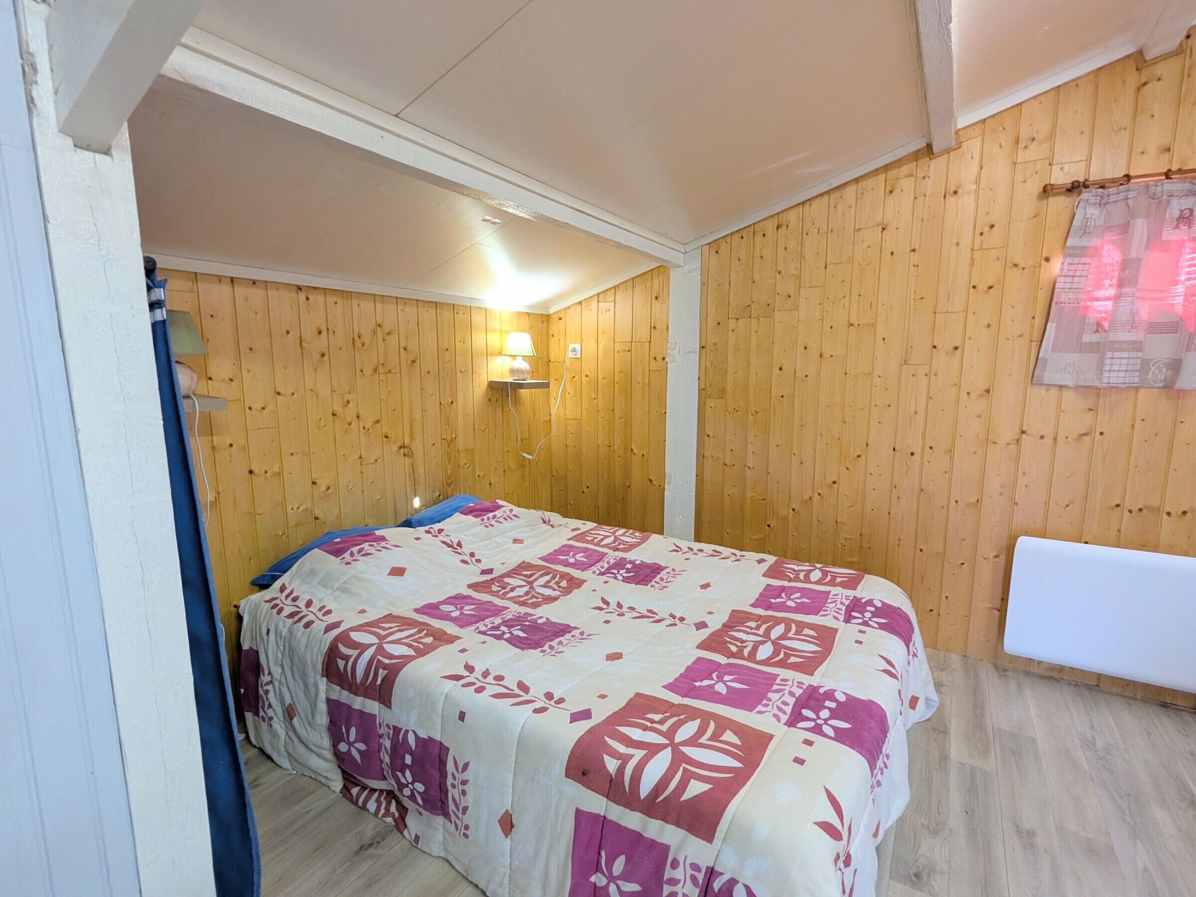 A bedroom with a bed, wooden walls, a wall-mounted lamp, and a window.