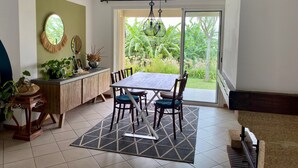 A dining area with a table and chairs, a kitchen counter with plants, a large window with a view of greenery, and a ceiling light fixture. - Cozy cottage, mountain view - 4 guests (Le Tampon)