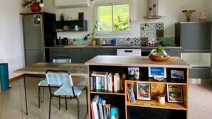 A modern kitchen with a dining area, a built-in bookshelf, and a window with a view of greenery. - Cozy cottage, mountain view - 4 guests (Le Tampon)