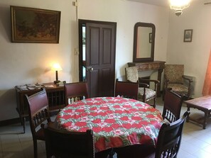 Une salle à manger avec une table ronde, une nappe à motifs floraux et des chaises en bois. On y trouve une porte, une chaise avec un coussin à motifs floraux et un miroir au mur.
