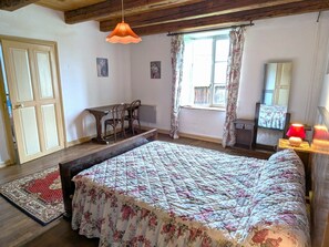 Une chambre à coucher avec un lit, un bureau et une chaise. Il y a une fenêtre avec des rideaux, un radiateur et un miroir.