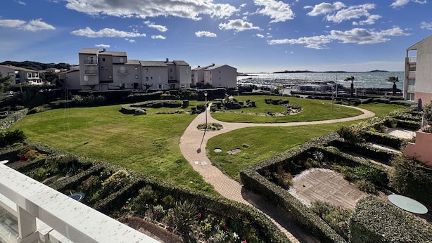 Un hotel sul lungomare con un cortile centrale, un porto turistico sullo sfondo e un cielo sereno.