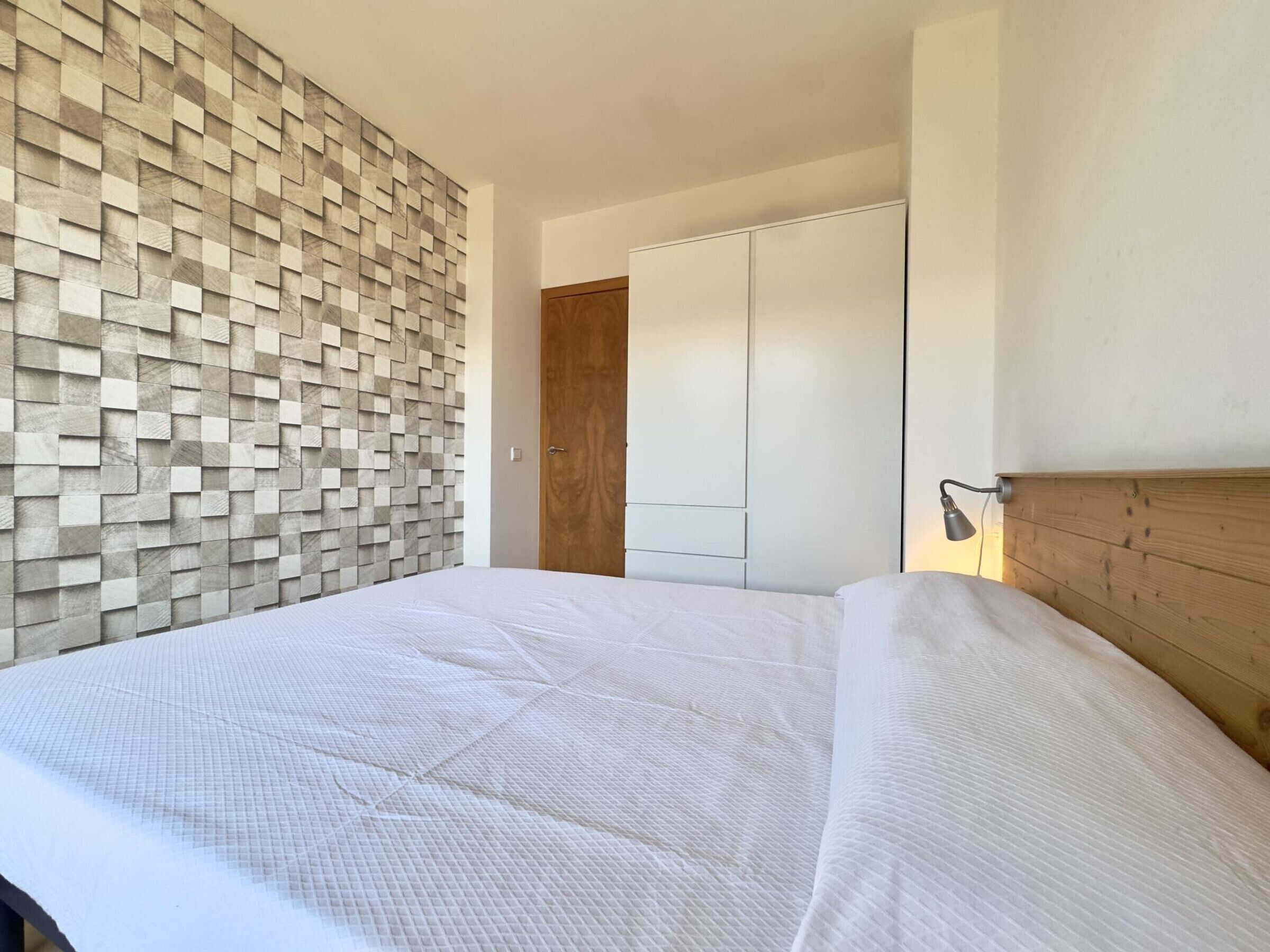 Une chambre à coucher avec un lit, une tête de lit en bois, un mur orné d’un motif géométrique et une armoire.