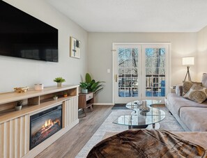 Una sala de estar con chimenea, un televisor de pantalla plana, una mesa de centro de vidrio, un sofá y una puerta que da al exterior.