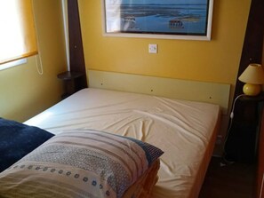 Un lit simple avec un matelas uni et un oreiller à motifs, une table de chevet avec une lampe, et un tableau représentant un paysage accroché au mur.