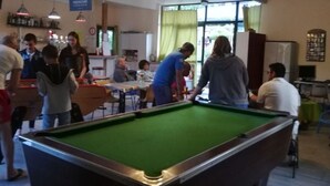 Une table de billard dans une pièce où se trouvent des tables et des chaises, un luminaire vert et une fenêtre avec des rideaux.