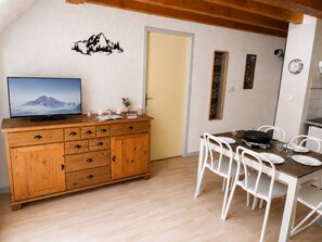 En matplats med ett träbord och vita stolar, ett träskåp med lådor, en tv på väggen och en dörr.