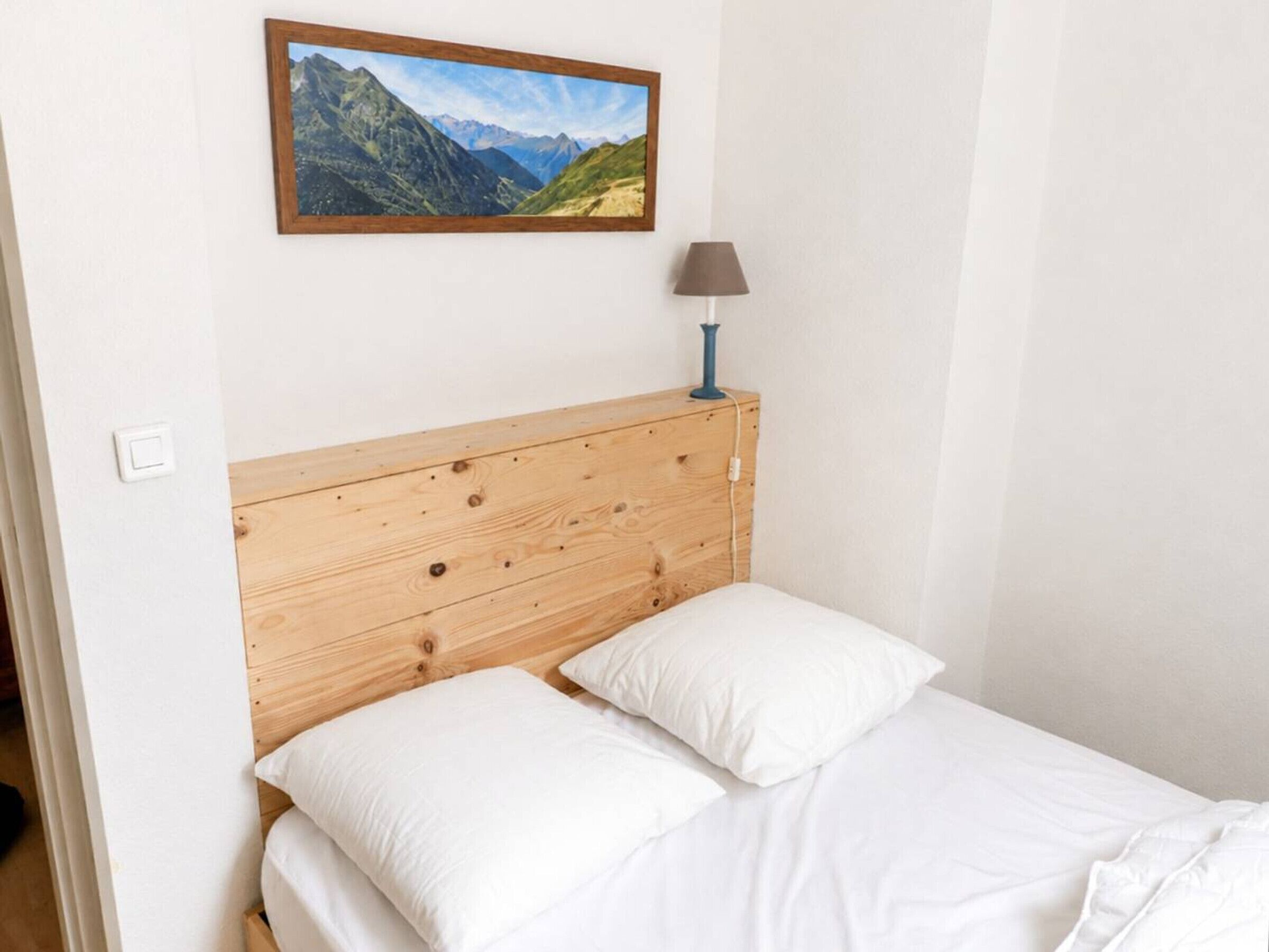A bedroom with a wooden headboard, white bedding, a lamp, and a mountain landscape painting on the wall.