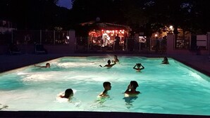 A swimming pool with people enjoying the water, surrounded by trees and a building with red lights in the background.