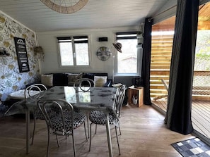 A dining area with a glass table and metal chairs, a wooden floor, and a view of the outdoors through large windows.
