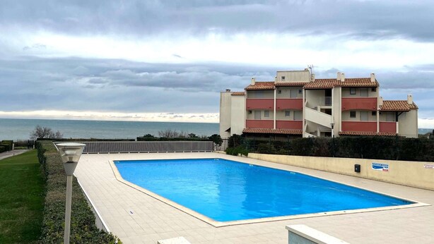 A swimming pool adjacent to a building with balconies, a view of the sea, and a cloudy sky. - Charming T2 sea view - 4 guests (Le CAP D'AGDE)