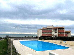 Une piscine située à côté d’un immeuble doté de balcons, avec vue sur la mer et sous un ciel nuageux.