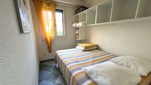 A small, tidy bedroom with a bed, a window with curtains, a shelf with storage boxes, and a tiled floor. - Charming T2 sea view - 4 guests (Le CAP D'AGDE)