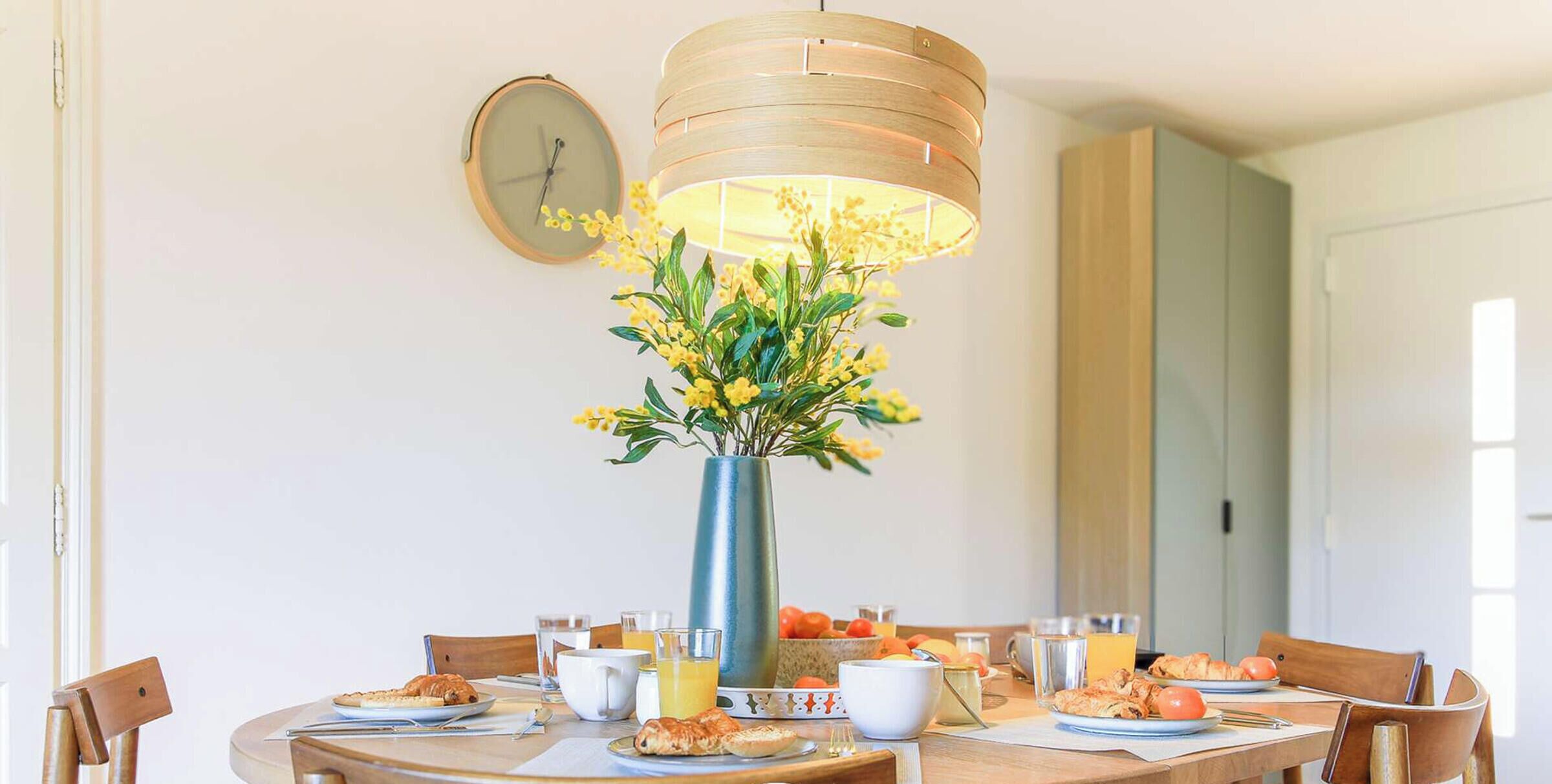 A dining table with breakfast items, a vase with yellow flowers, and a clock on the wall.