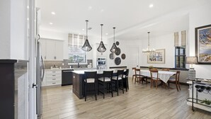 Una cocina moderna con armarios blancos, una isla central con sillas, iluminación colgante y un área de comedor con una mesa y sillas.