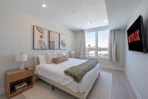 Ein modernes Schlafzimmer mit Bett, Nachttischen, einem Fernseher und Blick auf die Stadt.
