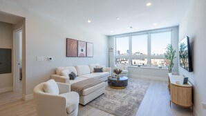 A modern living room with a large window, a sofa set, a coffee table, and a side table with a laptop. - Luxe 2BR Retreat 5-Min PATH to NYC With Rooftop+Gym (Jersey City)