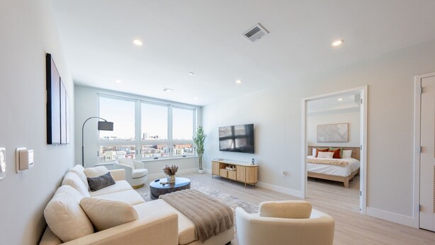 A modern living room with a large window, a flat-screen TV, a sofa, a coffee table, and a bed in the adjacent room. - Luxe 2BR Retreat 5-Min PATH to NYC With Rooftop+Gym (Jersey City)