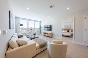 Un salon moderne doté d’une grande fenêtre, d’un téléviseur à écran plat, d’un canapé et d’une table basse, et une chambre adjacente avec un lit.