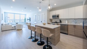 A modern kitchen with a central island, bar stools, and a large window offering a city view. - Luxe 2BR Retreat 5-Min PATH to NYC With Rooftop+Gym (Jersey City)