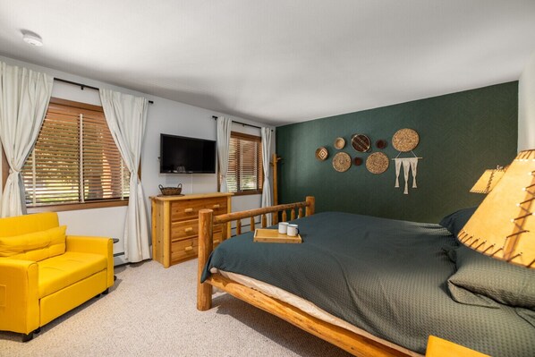 A bedroom with a bed, a wooden dresser, a yellow armchair, and a television on the wall.