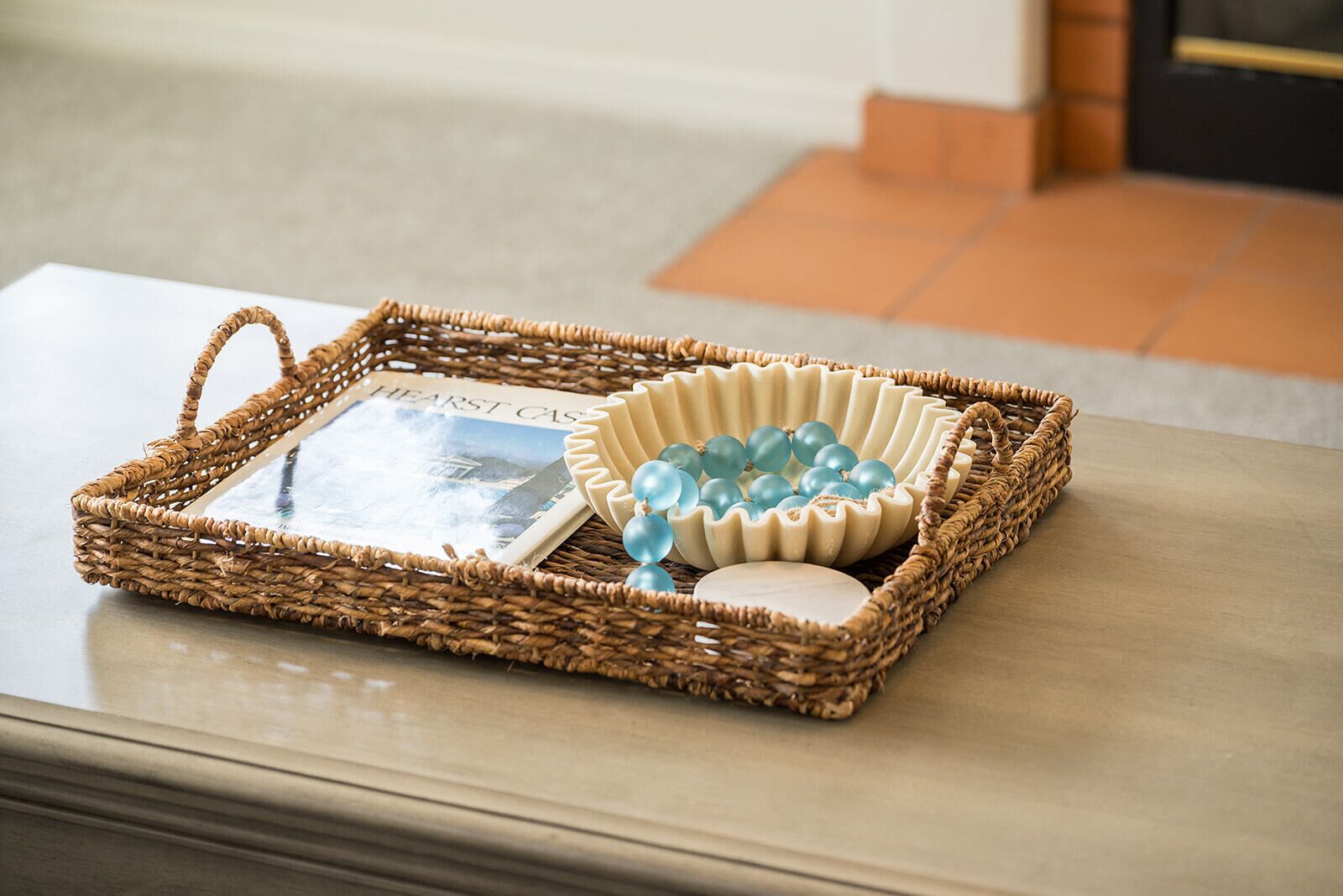 A wicker tray with a magazine, a soap dish, and a bowl of blue beads on a wooden table.
