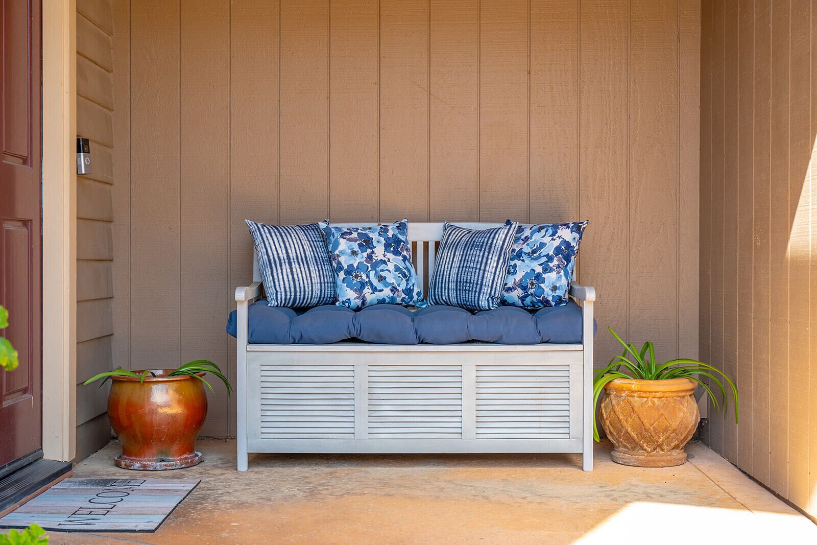 A white bench with blue cushions and patterned pillows, flanked by two large potted plants.