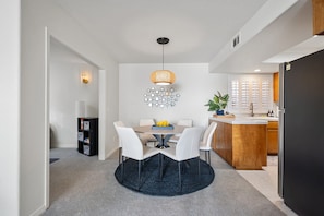A modern dining area with a round table, white chairs, a dark rug, a hanging light fixture, and a kitchen in the background.