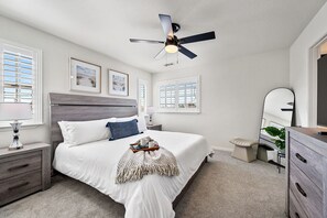 A bedroom with a bed, bedside tables, a nightstand, a ceiling fan, a dresser, a mirror, and a window with blinds.