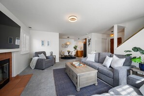 A modern living room with a fireplace, grey sofas, a coffee table, and a dining area in the background.