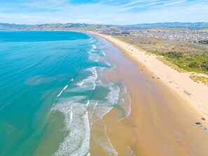 Une vue aérienne d’une plage aux eaux d’un bleu limpide, avec un rivage sablonneux et une silhouette de ville qui se dessine à l’horizon.