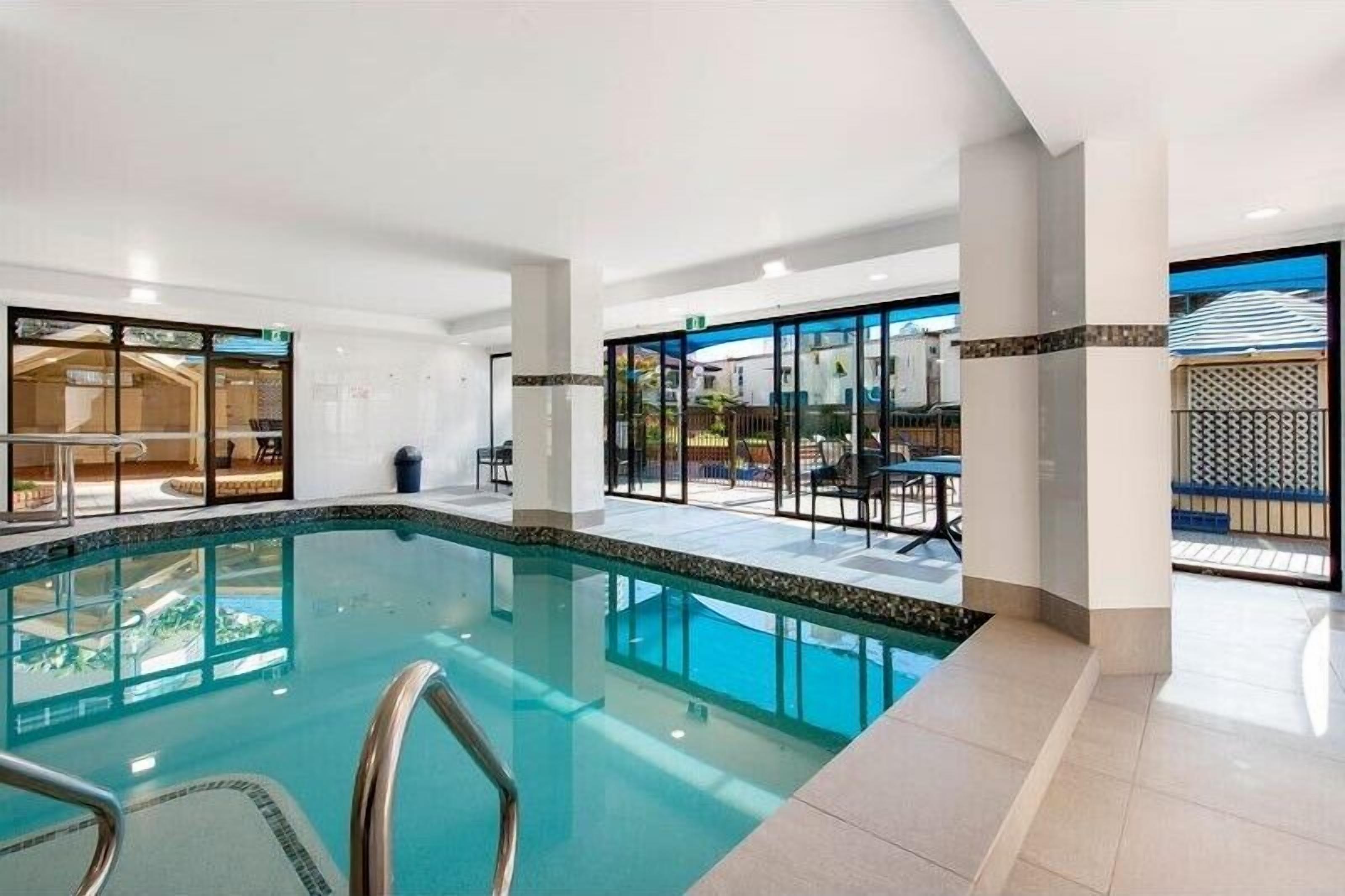 Piscina com água cristalina, rodeada por um deck revestido em azulejo e portas de vidro que dão para uma área externa com mesas e cadeiras.