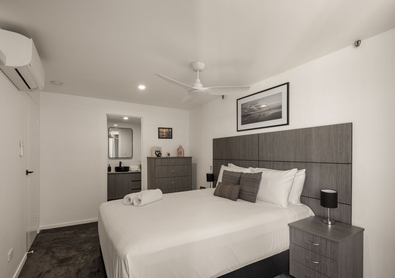 A modern bedroom with a large bed, a bedside table, a wardrobe, a mirror, and a ceiling fan.