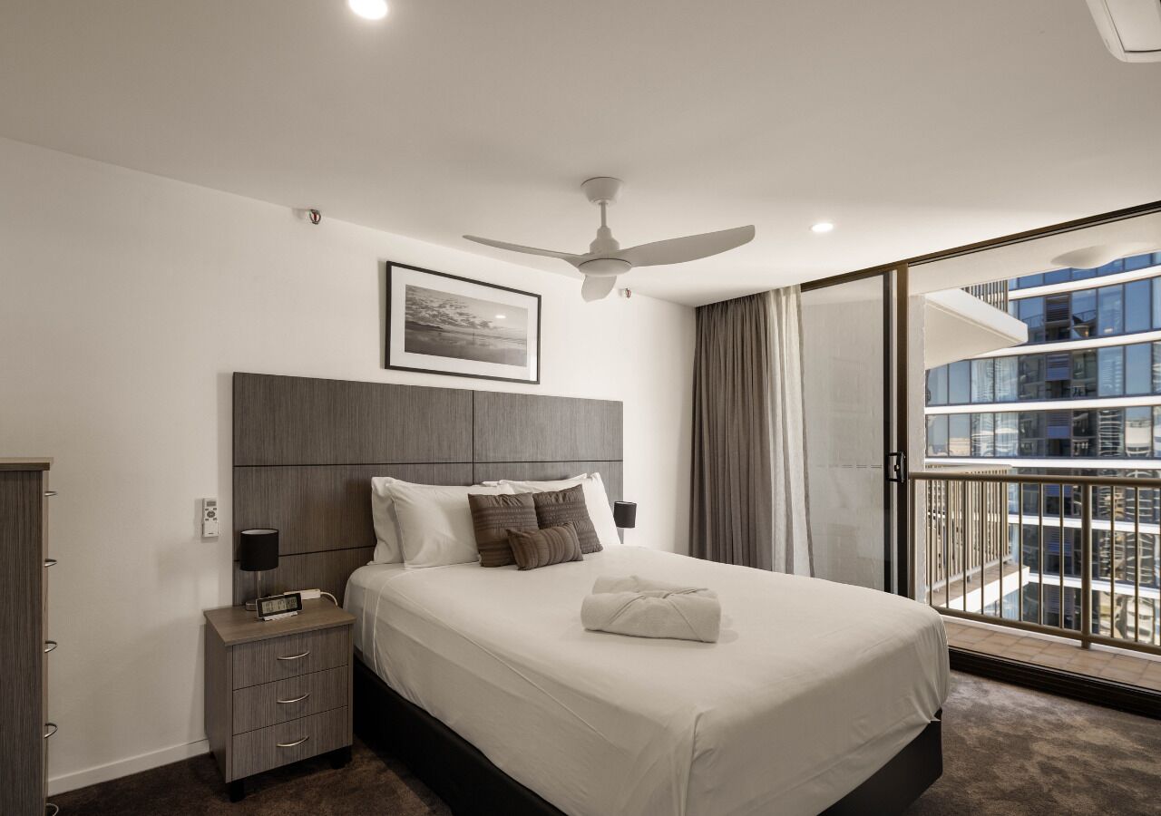 Ein modernes Hotelzimmer mit einem großen Bett, einem Nachttisch mit Lampe, einem Deckenventilator und Blick auf ein Gebäude durch eine Glastür.