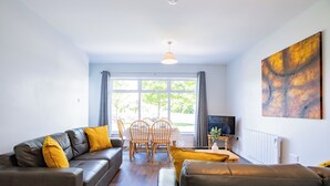 Ein modernes Wohnzimmer mit einer Couch, einem Couchtisch und einem Fernseher. Es gibt einen Esstisch mit Stühlen und ein Fenster mit Vorhängen.
