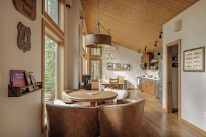 A spacious living area with a round table and leather chairs, wooden beams on the ceiling, and a kitchen in the background. - Roaring River Lodge- Modern Riverfront EV Fire Pit (Rhododendron)