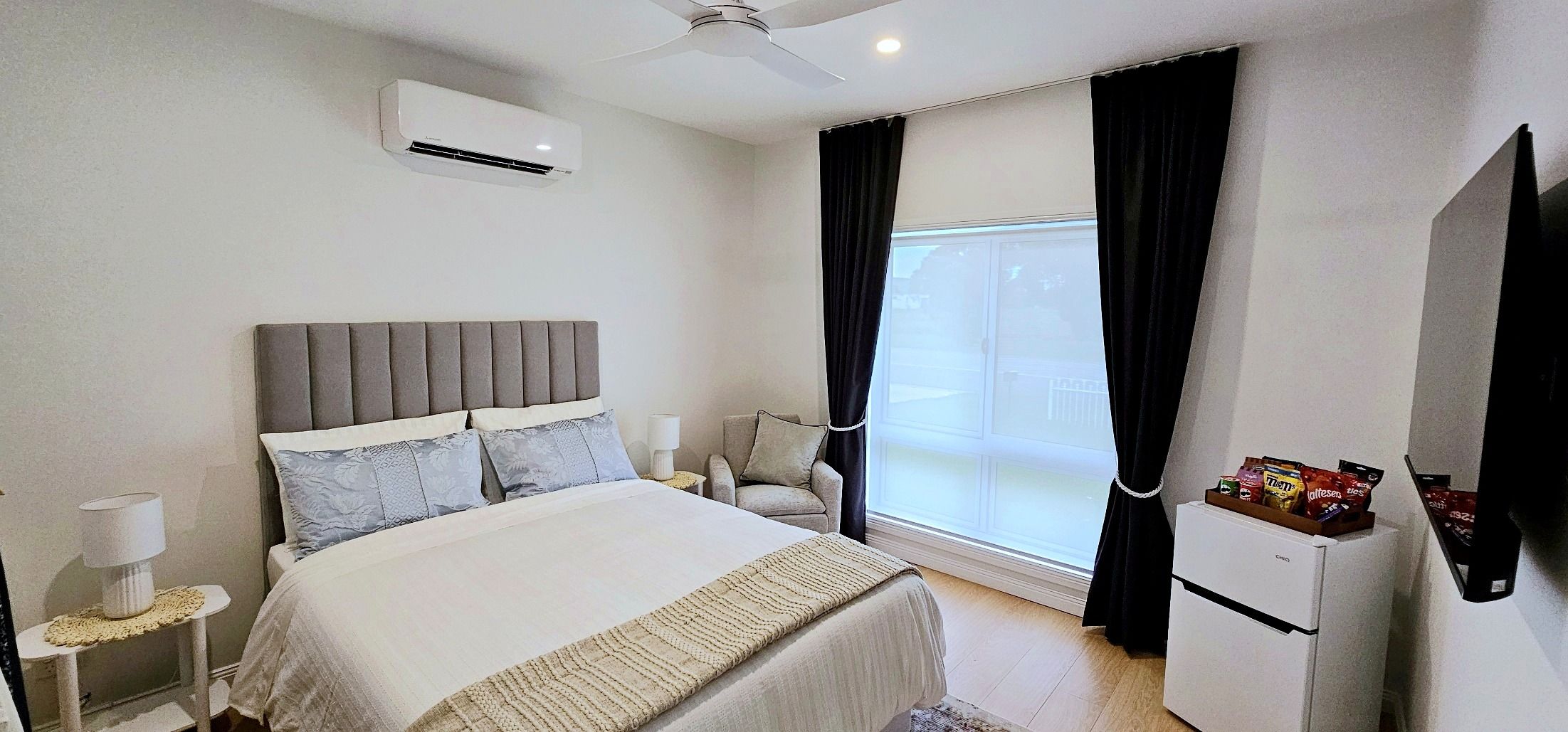 A hotel room with a bed, a chair, a desk, an air conditioner, a window with curtains, and a television.