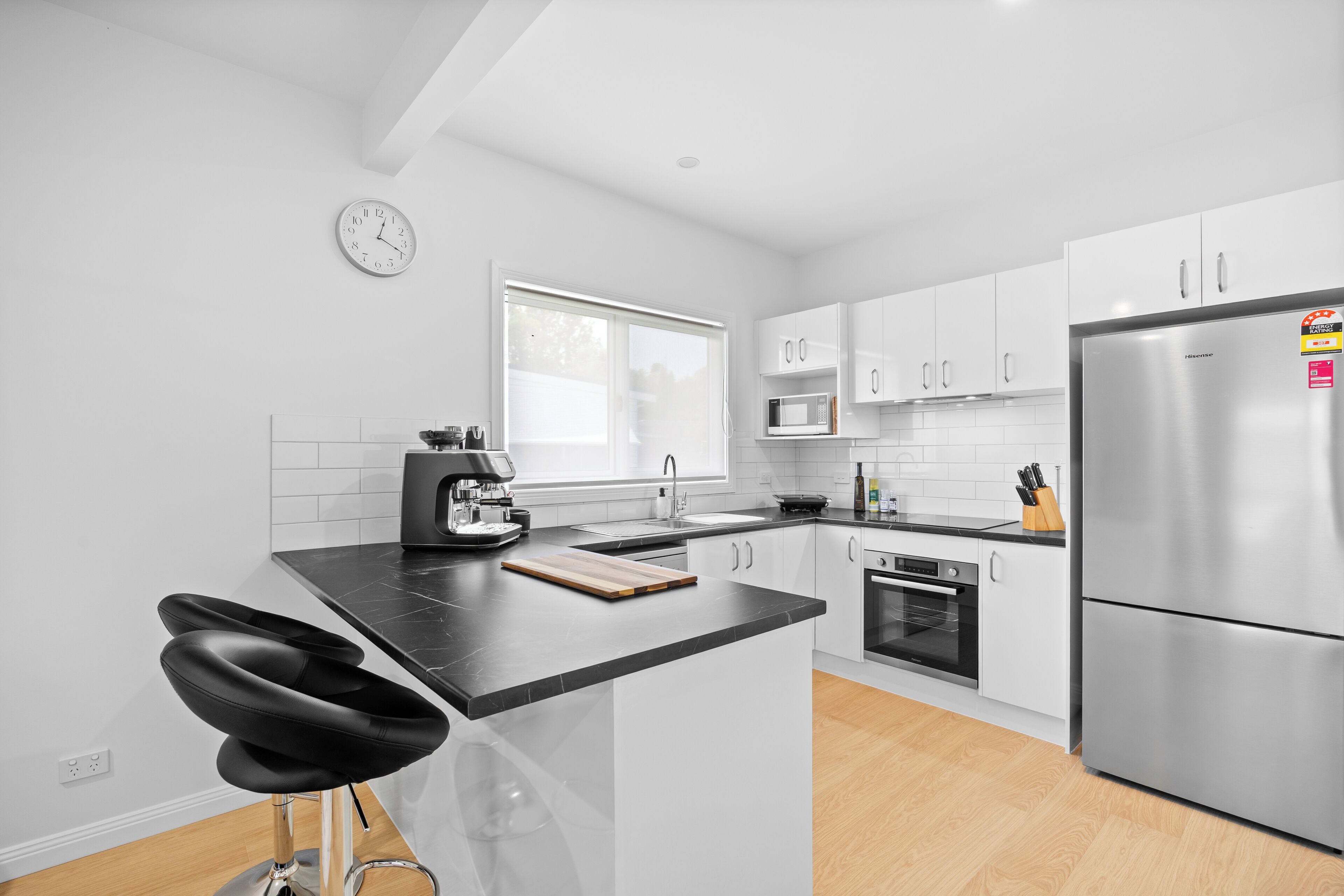 A modern kitchen with white cabinets, a stainless steel refrigerator, a coffee machine, and a window providing natural light.