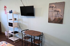 A room with a TV mounted on the wall, a wooden table with two chairs, a white shelf with various items, and a framed picture of birds. - BSI112--Studio with Shared Pool, Walk to Beach, Sleeps 2 Adults/2 Kids, Dogs OK! (Port Aransas)